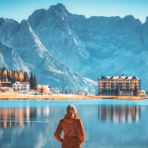 Young woman standing on the coast of Misurina lake at sunrise in autumn. Dolomites, Italy. Landscape with girl in red jacket, reflection in water, buildings, blue sky. Italian alps. Travel background